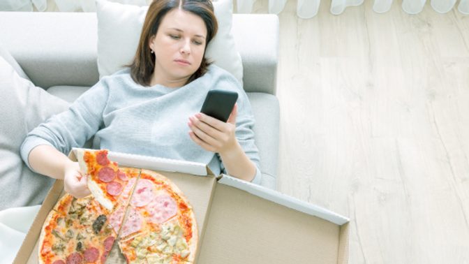 A woman lies on the couch eating a whole pizza while looking at her phone