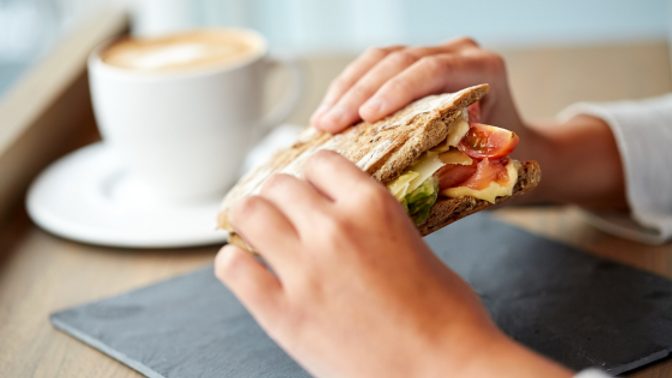 Hands hold a sandwich in front of a cup of coffee