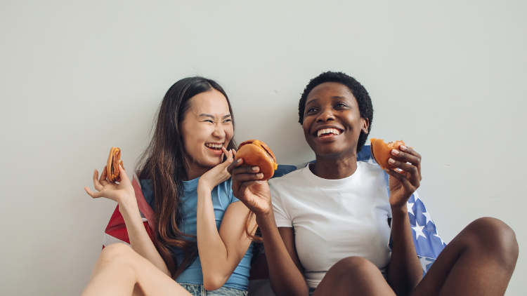 Two women sit against a wall eating cheeseburgers as part of their intuitive eating practice